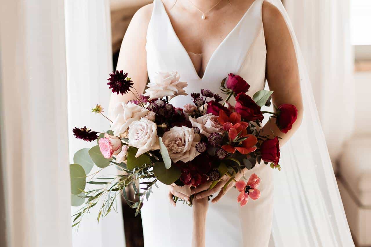 Josie Blair - Wedding Day Tuscany Italy A bride in a white dress holds a bouquet with a mix of red, pink, and cream flowers, along with green leaves. The image captures the upper part of the bride, emphasizing the bouquet and the dress's neckline.
