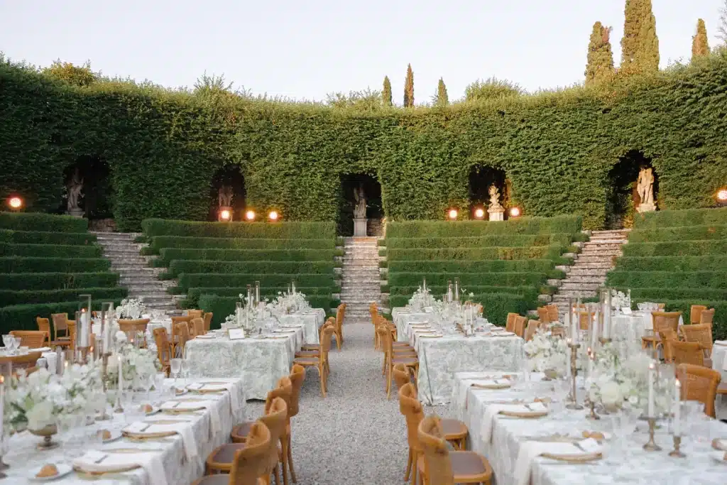 An outdoor formal dining setup at Villa Rizzardi features long tables decorated with white tablecloths, floral centerpieces, and candle holders. The area is surrounded by lush green hedges, steps, and statues, creating an elegant garden atmosphere. Rows of chairs are arranged neatly around the tables.