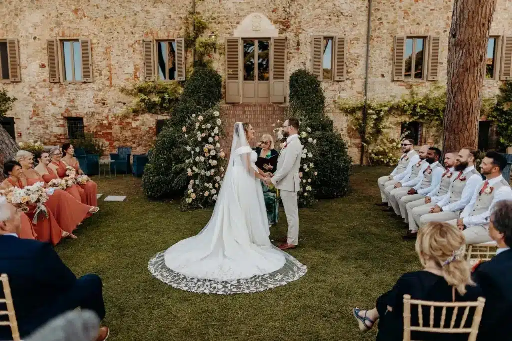 Leslie and Wayne's Tuscan Wedding. A bride and groom stand at the altar during an outdoor wedding ceremony. The bride wears a long white dress, and the groom wears a light suit. Guests sit on either side, with the ceremony set against a rustic stone building with greenery.