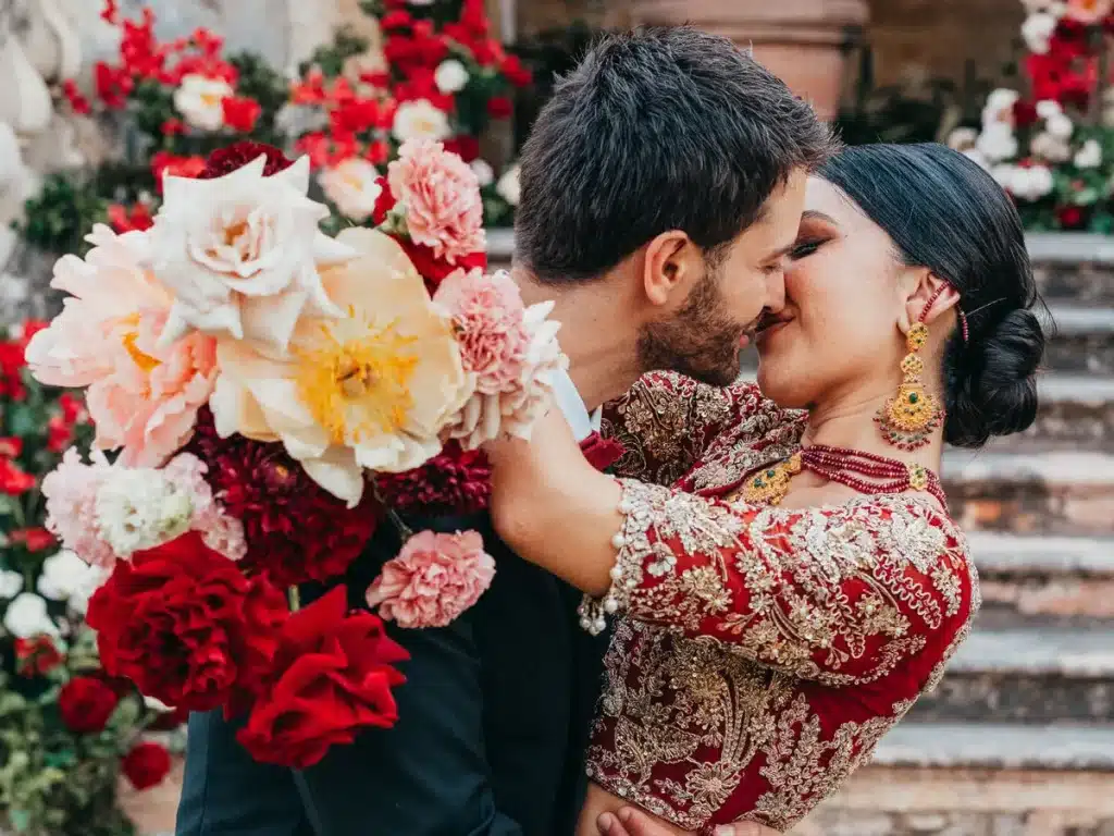 Wedding at Villa Arvedi. A couple embraces and kisses passionately on a stone staircase, surrounded by colorful flowers. The woman wears an ornate red and gold outfit, with matching jewelry, while holding a vibrant bouquet of red, white, and yellow flowers.
